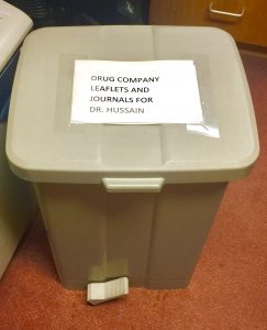 A grey pedal bin in a consulting room with a printed label reading Drug Company Leaflets And Journals For Dr. Hussain